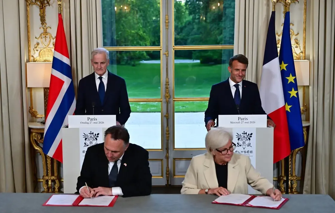 Two ministers sign documents at a table in an Élysée salon while Jonas Gahr Støre and Emmanuel Macron stand behind them at lecterns, between Norwegian, French and EU flags.